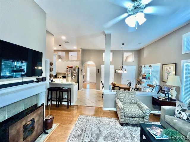 living area featuring a tiled fireplace, arched walkways, baseboards, ceiling fan with notable chandelier, and light wood-style floors