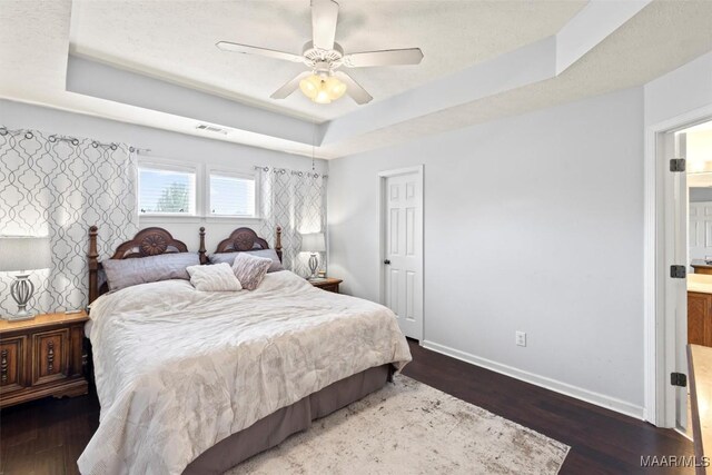 bedroom featuring a tray ceiling, wood finished floors, baseboards, ceiling fan, and ensuite bath