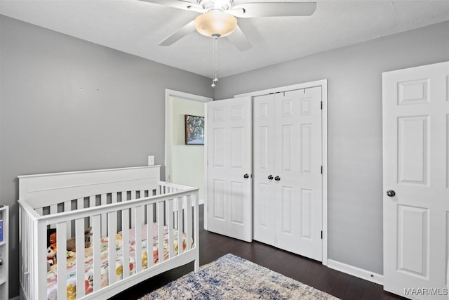 bedroom with a closet, dark wood-type flooring, a nursery area, baseboards, and ceiling fan