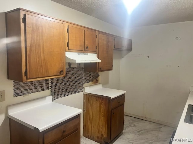 kitchen with under cabinet range hood, light marble finish floors, backsplash, light countertops, and brown cabinetry