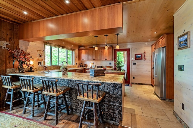 kitchen with kitchen peninsula, a breakfast bar, wooden walls, wooden ceiling, and stainless steel refrigerator