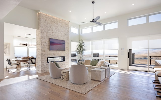 living room featuring ceiling fan, a towering ceiling, a stone fireplace, and light wood-type flooring