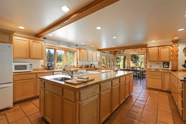 kitchen with an island with sink, white appliances, sink, light tile floors, and beamed ceiling