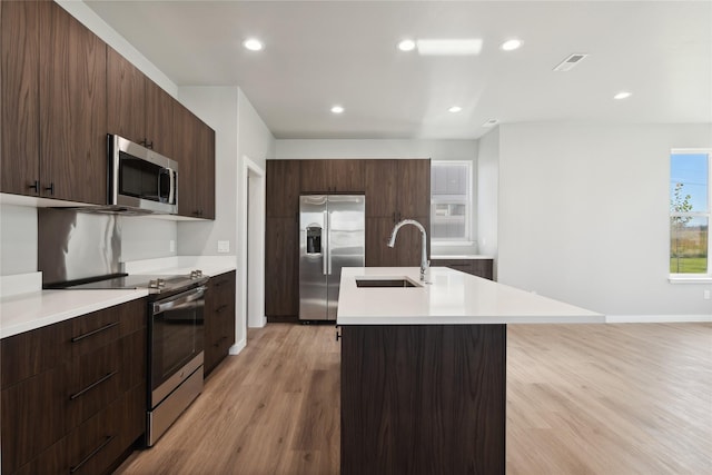 kitchen with sink, dark brown cabinets, stainless steel appliances, a center island with sink, and light wood-type flooring