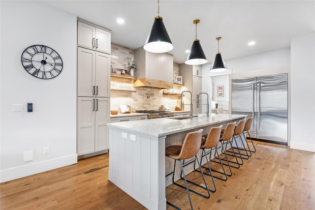 kitchen featuring light stone countertops, stainless steel built in fridge, decorative backsplash, hanging light fixtures, and a kitchen island with sink