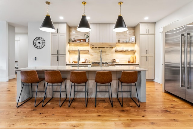 kitchen with built in refrigerator, hanging light fixtures, an island with sink, and light stone counters