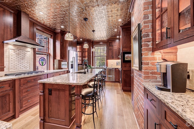 kitchen with a breakfast bar, light hardwood / wood-style flooring, appliances with stainless steel finishes, wall chimney range hood, and a center island with sink