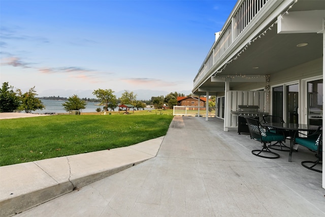 patio terrace at dusk with grilling area, a water view, and a lawn