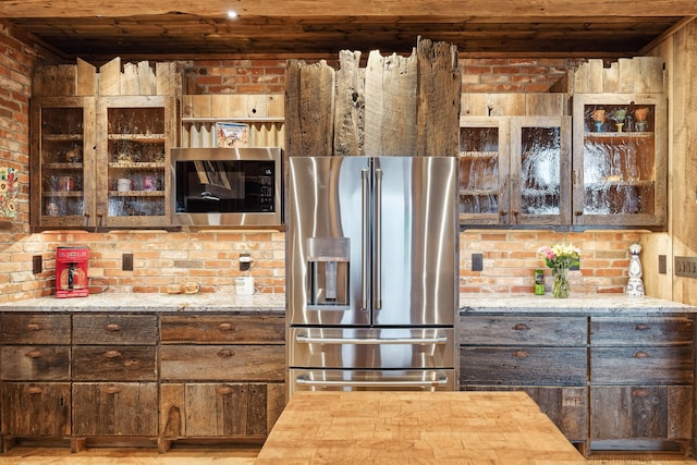 kitchen with dark brown cabinetry, light stone countertops, stainless steel appliances, and brick wall