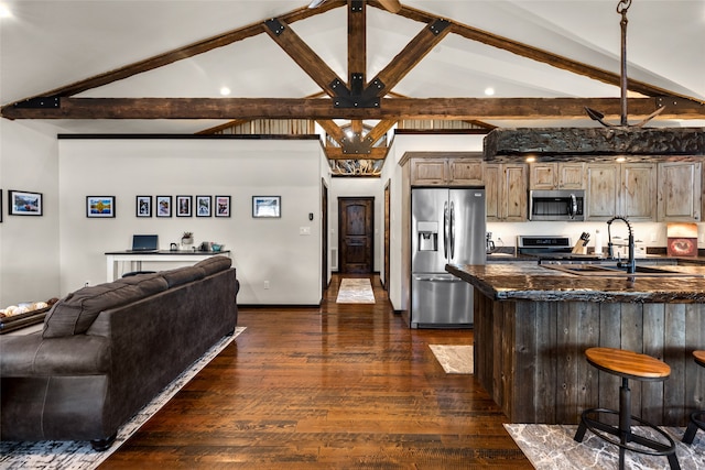 kitchen featuring dark stone counters, appliances with stainless steel finishes, beamed ceiling, dark hardwood / wood-style floors, and a kitchen bar