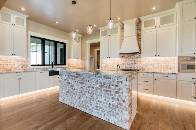 1052 Foxtail Dr, Kalispell, MT 59901 - kitchen featuring an island with sink, stainless steel appliances, light wood finished floors, premium range hood, and light stone countertops