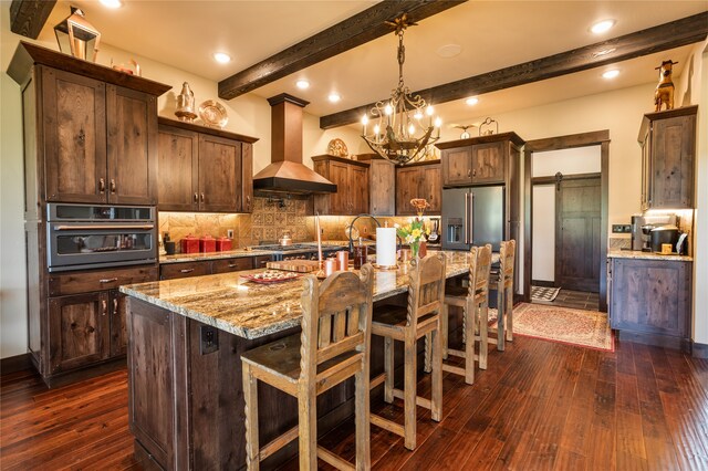 kitchen featuring stainless steel appliances, beam ceiling, wall chimney range hood, a kitchen island with sink, and dark hardwood / wood-style flooring