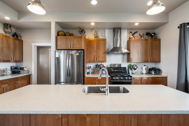 kitchen with gas stove, sink, stainless steel refrigerator, wall chimney exhaust hood, and a center island with sink