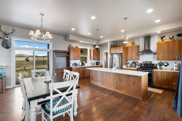 kitchen featuring stainless steel fridge, decorative light fixtures, a center island with sink, wall chimney exhaust hood, and black range oven