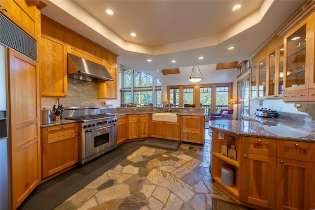 kitchen with stone countertops, high end stove, a peninsula, wall chimney exhaust hood, and a raised ceiling