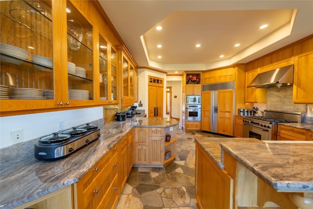 kitchen featuring a raised ceiling, glass insert cabinets, light stone counters, built in appliances, and wall chimney range hood