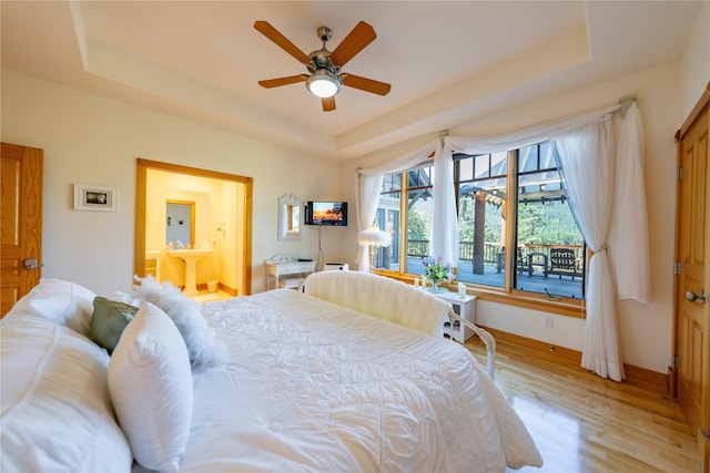 bedroom with a tray ceiling, light wood-type flooring, connected bathroom, and a ceiling fan