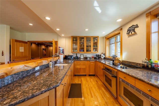 kitchen with stainless steel appliances, a peninsula, a sink, dark stone counters, and glass insert cabinets