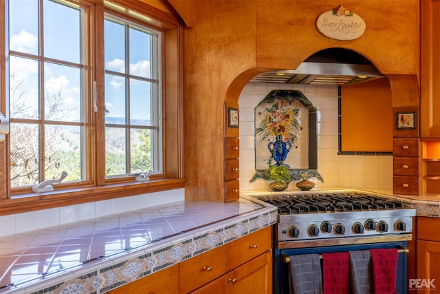 kitchen featuring wall chimney exhaust hood, tile countertops, stainless steel stove, and backsplash