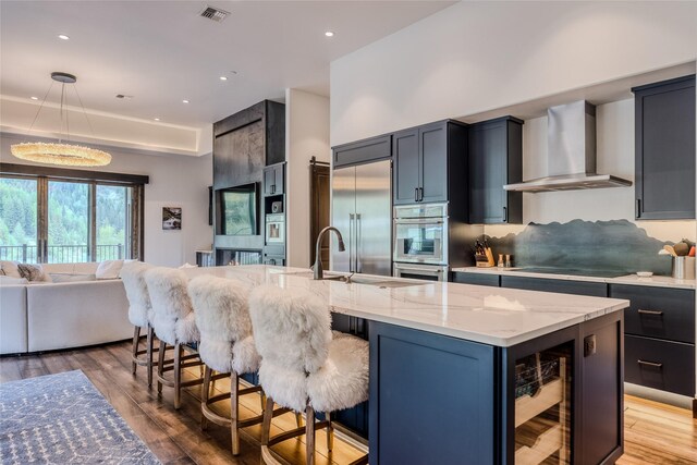 kitchen with pendant lighting, wall chimney range hood, stainless steel appliances, and a kitchen island with sink