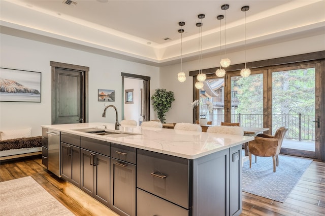 kitchen featuring a tray ceiling, a kitchen island with sink, hanging light fixtures, light stone counters, and sink