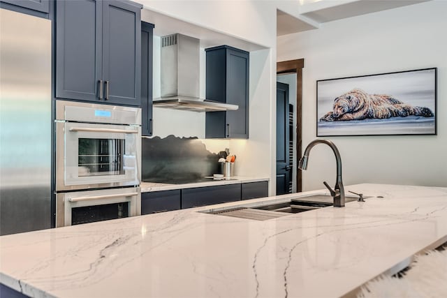 kitchen featuring light stone countertops, wall chimney range hood, stainless steel appliances, sink, and blue cabinetry