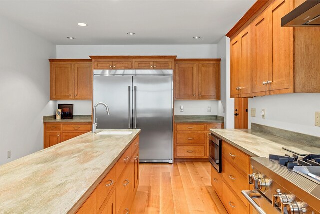 kitchen with a center island with sink, light wood-type flooring, wall chimney range hood, high end appliances, and sink