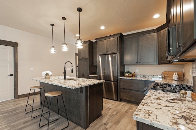 kitchen with decorative light fixtures, sink, a center island with sink, stainless steel fridge, and light hardwood / wood-style flooring