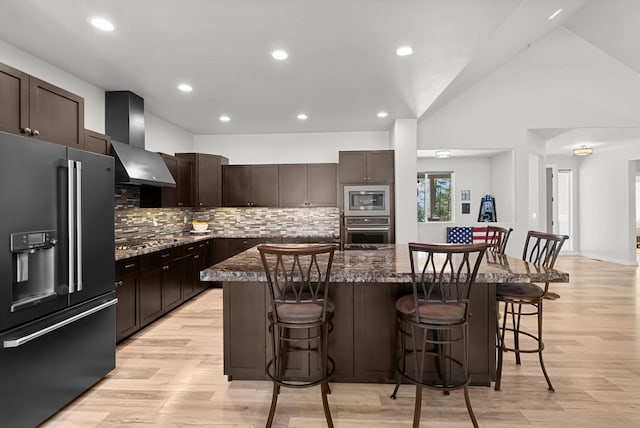 kitchen with wall chimney range hood, appliances with stainless steel finishes, a breakfast bar area, and an island with sink