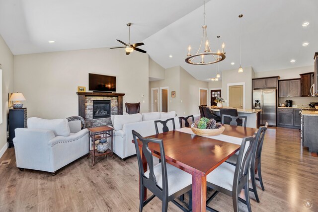 dining room with ceiling fan with notable chandelier, wood-type flooring, a fireplace, and vaulted ceiling