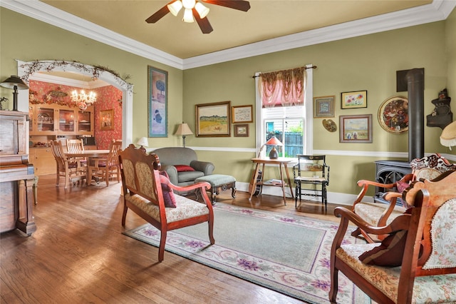 sitting room with wood-type flooring, ceiling fan with notable chandelier, a wood stove, and ornamental molding