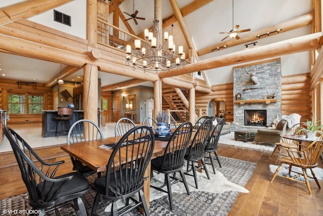 dining area featuring high vaulted ceiling, ceiling fan with notable chandelier, a stone fireplace, log walls, and wood-type flooring