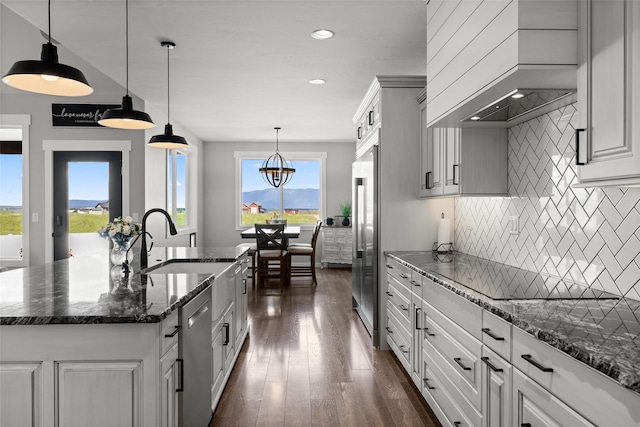 kitchen with high quality fridge, custom range hood, pendant lighting, a kitchen island with sink, and decorative backsplash