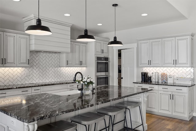 kitchen with a kitchen island with sink, double oven, decorative light fixtures, and a kitchen bar