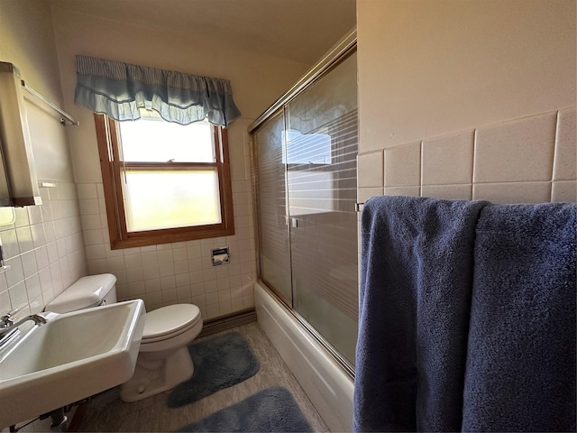 bathroom featuring toilet, bath / shower combo with glass door, a sink, and tile walls