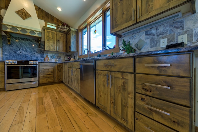 kitchen featuring custom exhaust hood, stainless steel appliances, decorative backsplash, vaulted ceiling, and light hardwood / wood-style flooring