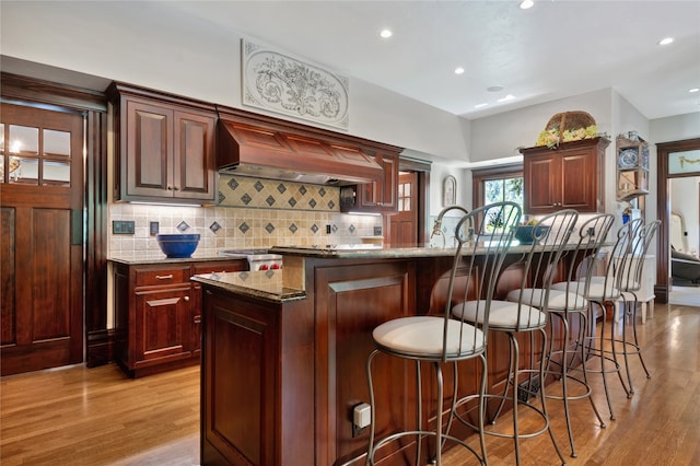 kitchen featuring wall chimney range hood, light wood-type flooring, light stone counters, and an island with sink