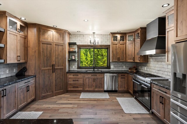 kitchen featuring backsplash, dark hardwood / wood-style floors, wall chimney range hood, dark stone countertops, and appliances with stainless steel finishes