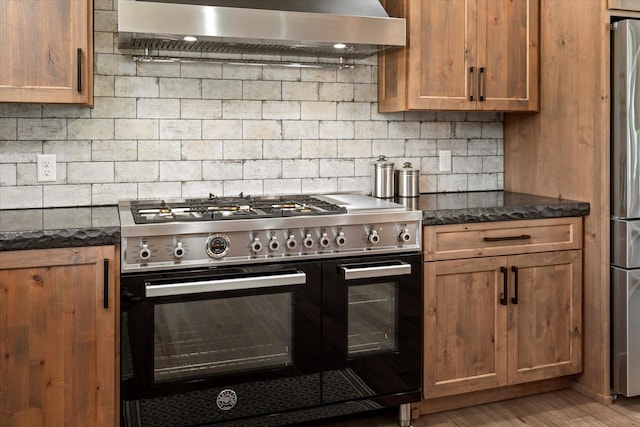 kitchen featuring appliances with stainless steel finishes, dark stone counters, decorative backsplash, light hardwood / wood-style floors, and range hood