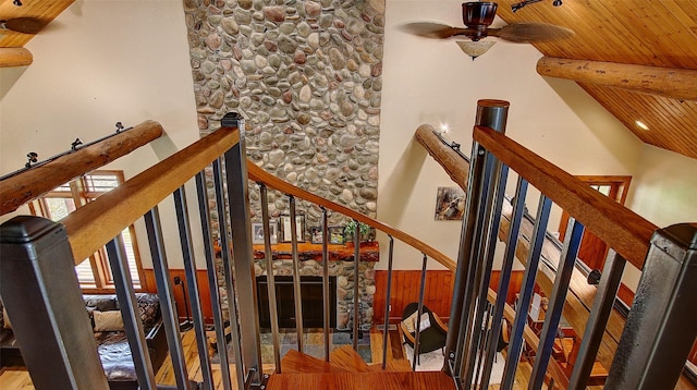 stairway with ceiling fan, a stone fireplace, vaulted ceiling with beams, wooden walls, and wood ceiling