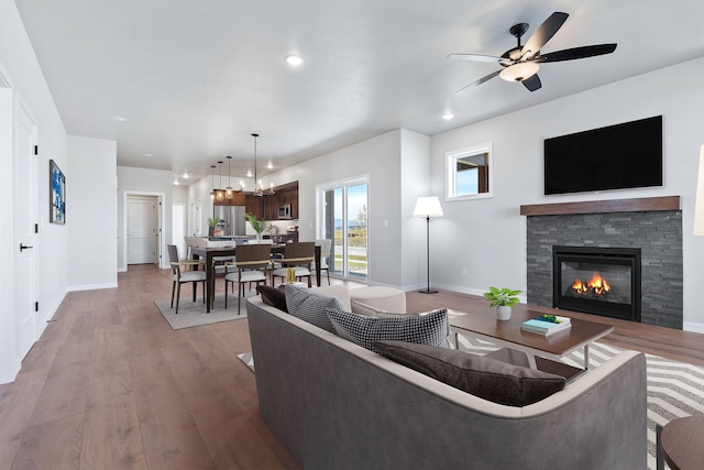 living room featuring light hardwood / wood-style flooring, ceiling fan with notable chandelier, and a stone fireplace