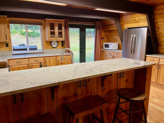 kitchen featuring light wood-type flooring, light stone countertops, stainless steel refrigerator, beamed ceiling, and wood walls