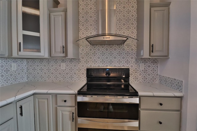 kitchen with glass insert cabinets, light stone countertops, stainless steel range with electric stovetop, wall chimney range hood, and backsplash