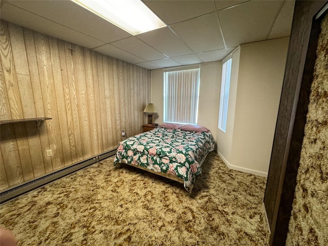 carpeted bedroom featuring a drop ceiling, baseboard heating, and wood walls