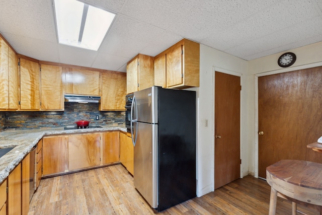 kitchen featuring freestanding refrigerator, light wood-style flooring, black electric cooktop, light stone counters, and under cabinet range hood