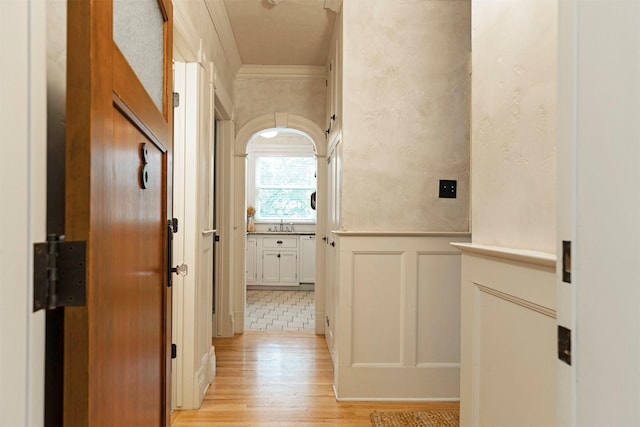 corridor with light wood-type flooring, a wainscoted wall, ornamental molding, and a decorative wall