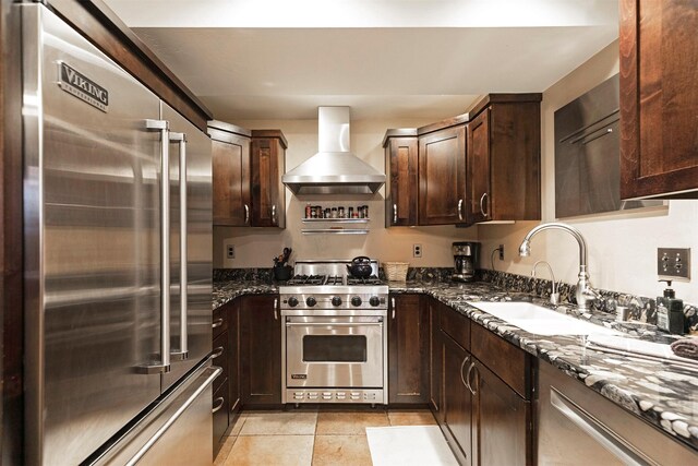 kitchen with dark brown cabinets, light tile patterned floors, wall chimney range hood, dark stone counters, and high quality appliances