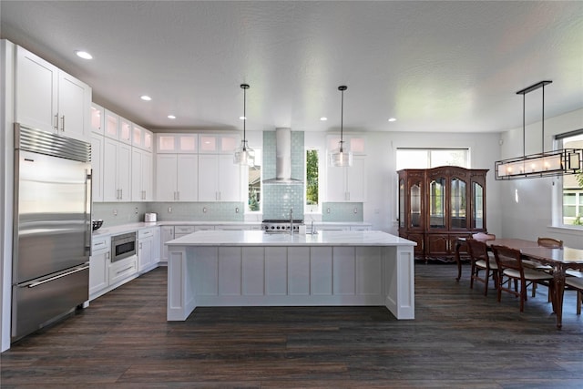 kitchen with a center island with sink, wall chimney range hood, dark wood finished floors, light countertops, and built in appliances