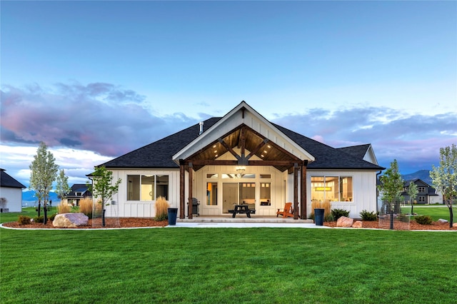 back of house at dusk featuring a lawn, board and batten siding, a shingled roof, and a patio