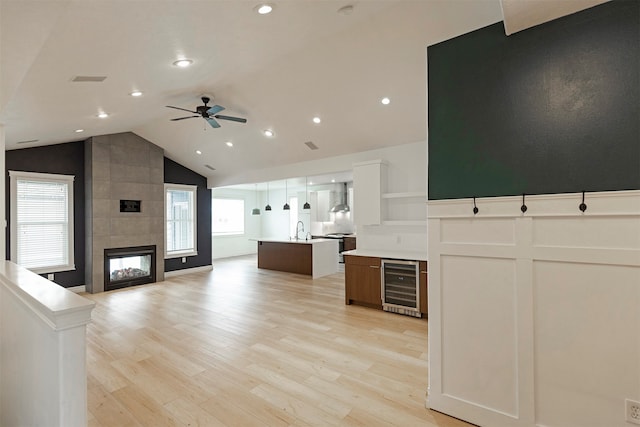 kitchen featuring a center island with sink, wine cooler, wall chimney exhaust hood, ceiling fan, and a tiled fireplace